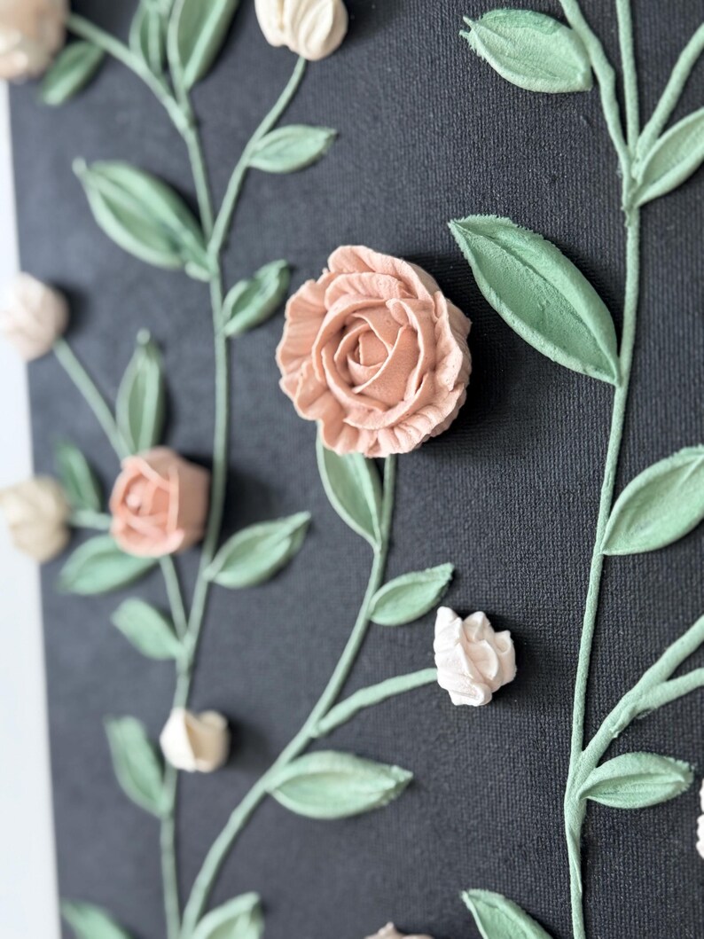 May include: Close-up of a canvas artwork with raised floral elements. The piece features peach roses and off-white buds, with green leaves and stems against a dark grey background. The textured design creates a three-dimensional effect.