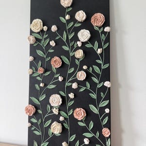 May include: A black rectangular canvas with a floral design. Raised, textured roses in cream and peach tones are arranged with green stems and leaves. The artwork is mounted on a wooden surface, creating a 3D effect.