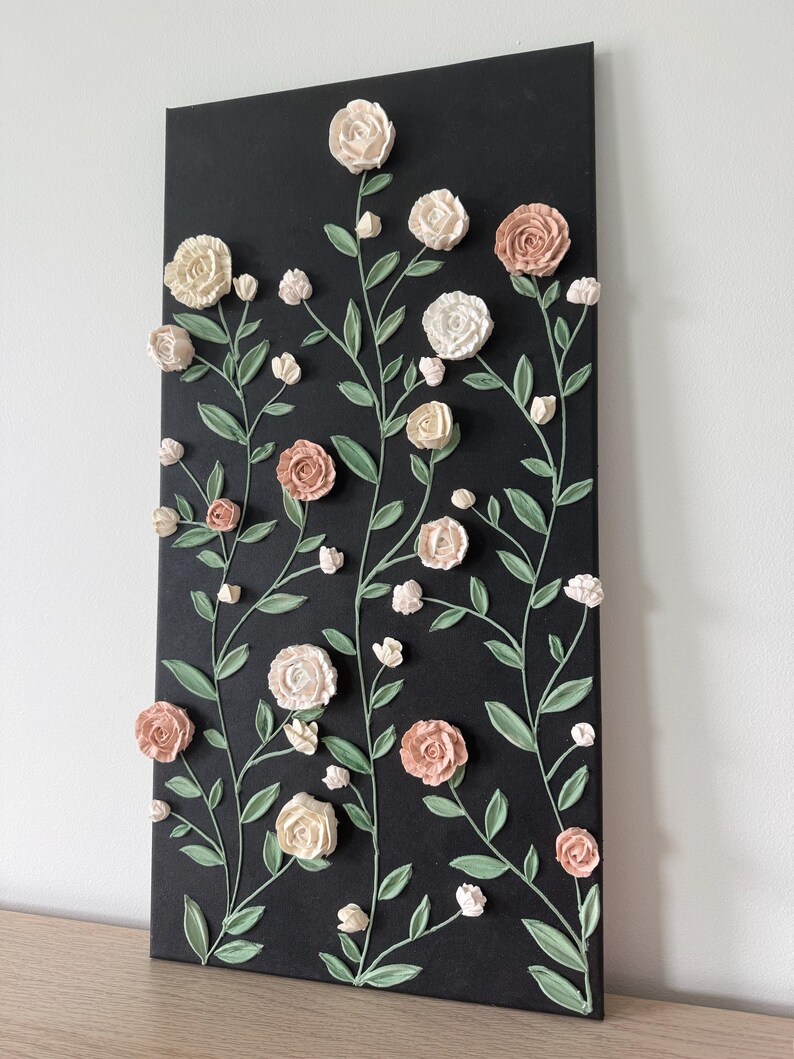 May include: A black rectangular canvas with a floral design. Raised, textured roses in cream and peach tones are arranged with green stems and leaves. The artwork is mounted on a wooden surface, creating a 3D effect.