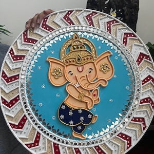 May include: A decorative round wall hanging featuring a cartoon depiction of the Hindu deity Ganesha. The artwork has a light blue background with a central figure of Ganesha in orange and blue. The border is white, red, and beige with mirrored accents.