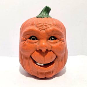 May include: Orange ceramic pumpkin with a smiling face, black eyes, and a green stem. The pumpkin has a textured surface and a single tooth. A Halloween decoration.