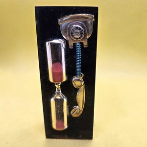 May include: A black rectangular decorative piece featuring a gold-colored vintage telephone and an hourglass with red sand. The telephone has a blue coiled cord and a handset. The hourglass is made of clear glass.