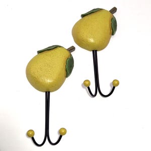 May include: Two decorative wall hooks shaped like pears. The pear-shaped tops are yellow with green leaves and brown stems. The hooks are black metal with yellow ball ends, designed for hanging items. A charming and functional home decor item.
