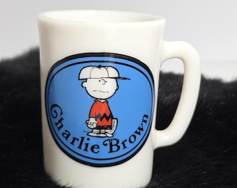 May include: A white ceramic mug featuring a blue oval graphic on the front. Inside the oval is an illustration of the character Charlie Brown wearing a white cap and an orange shirt with a black zigzag pattern. The text Charlie Brown is printed.