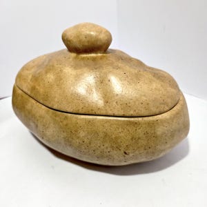 May include: A tan ceramic lidded container with a rounded, organic shape. The lid has a small, rounded knob. The container has a speckled texture and a matte finish, suggesting a handmade or rustic aesthetic. The overall design is simple and functional.