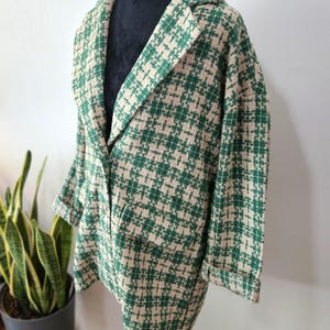 May include: A green and beige houndstooth pattern coat with a notched collar and two front pockets. The coat is displayed on a black mannequin and has rolled-up sleeves. The coat is made of a textured fabric.