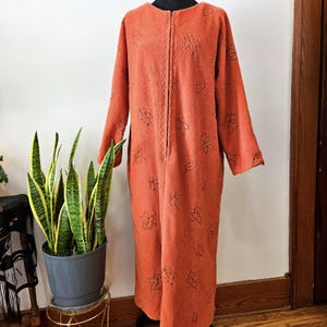 May include: An orange, long-sleeved robe with a full-length zipper and leaf pattern. The robe is made of a soft, textured material and is displayed on a mannequin. The leaves are black and the zipper has a scalloped edge.