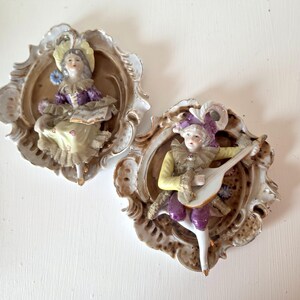 May include: Two antique porcelain wall plaques featuring figures in period costumes. One figure is seated and reading, the other is playing a lute. Both are set in ornate, gilded frames with a cream and brown color scheme.