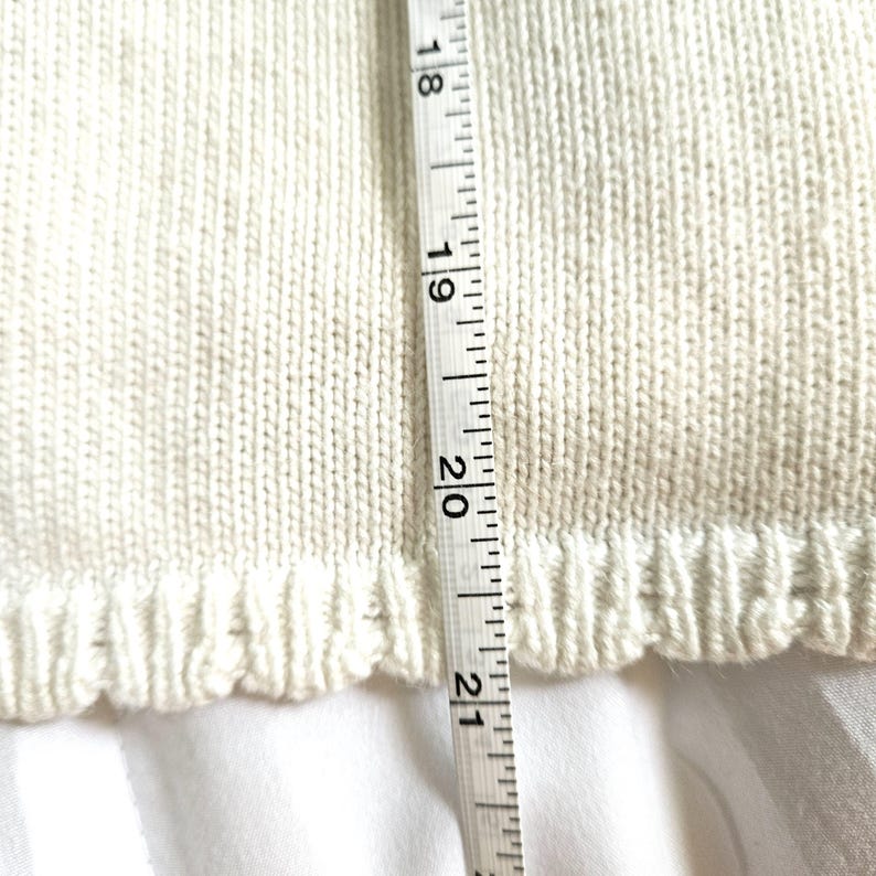 May include: Close-up of a cream-coloured knitted garment with a scalloped edge. A measuring tape shows a length of approximately 45.7 to 53.3 cm. The fabric has a ribbed texture.