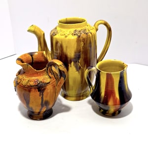 May include: A collection of ceramic kitchenware in shades of yellow, brown, and orange. The set includes a tall teapot, a small pitcher, and a creamer. The teapot has a decorative floral band around the top. The pitcher and creamer have handles.