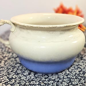 May include: A cream-colored ceramic pot with a blue base and a decorative handle. The pot has a wide opening and a slightly distressed finish, suggesting an antique or vintage style. The pot is likely used for decorative purposes.