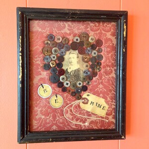 May include: Framed artwork featuring a vintage portrait at the center, surrounded by a heart-shaped arrangement of assorted buttons. The background is a red patterned fabric. Includes tags that read "B", "E", and "MINE". The frame is black with gold accents.