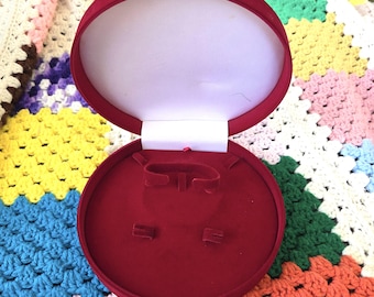May include: Open, round, burgundy jewelry box with a white interior and a hinged lid. The box has a velvet-like texture and features small holders for earrings and a necklace. The box is on a colorful, patterned surface.