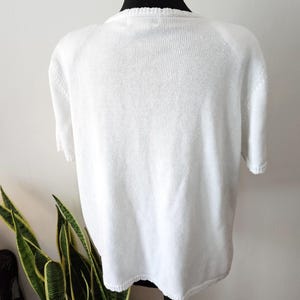 May include: A white short-sleeved knitted top. The top is displayed on a black mannequin. The knitted fabric has a textured appearance. The neckline and sleeves have a ribbed design. The top is a solid white colour.