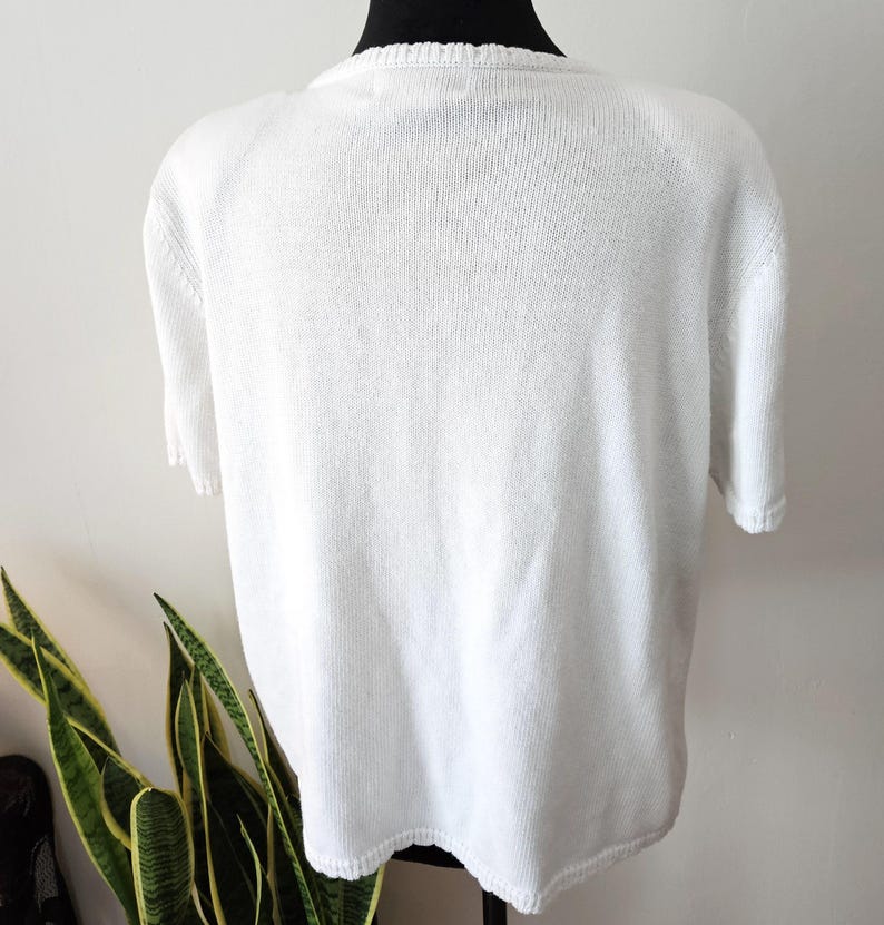 May include: A white short-sleeved knitted top. The top is displayed on a black mannequin. The knitted fabric has a textured appearance. The neckline and sleeves have a ribbed design. The top is a solid white colour.