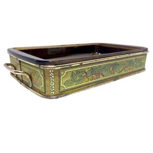 May include: A rectangular metal tray with a dark brown interior and a gold-toned exterior featuring a decorative border of green leaves and orange fruit. The tray includes two silver-colored metal handles on the sides and a geometric pattern.
