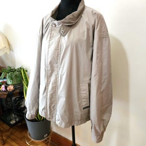 May include: A tan jacket with a button-up collar and a single button closure. The jacket has a front pocket and a small logo on the pocket. The jacket is displayed on a black mannequin.