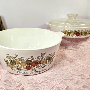 May include: Two white ceramic casserole dishes with lids. The dishes feature a floral pattern with a variety of vegetables and herbs. The text "L'Echalote La Marjolaine" is printed on the bottom of the dish.