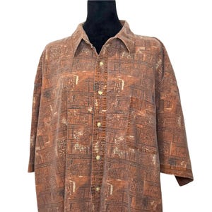 May include: A short-sleeved button-down shirt in a muted rust-brown color featuring an all-over geometric pattern of dark brown and cream-colored rectangular shapes. The shirt has a standard collar, a front button placket, and a chest pocket.
