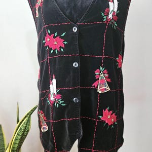 May include: Black velvet vest with a V-neck and button closure. The vest features embroidered Christmas-themed designs, including poinsettias, candles, and bells, outlined with red stitching. The vest has a grid pattern.