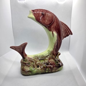 May include: A ceramic fish-shaped decorative item. The fish is primarily burgundy with a light green underbelly and base. The base has several small holes. The fish is arched, with its mouth open. The tail fin is visible.