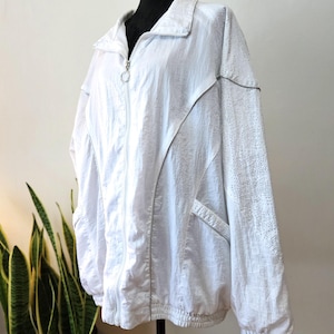 May include: White athletic jacket with a zip-up front, collar, and elasticized cuffs and hem. The jacket features silver trim and perforated detailing on the sleeves. The jacket is displayed on a black mannequin.