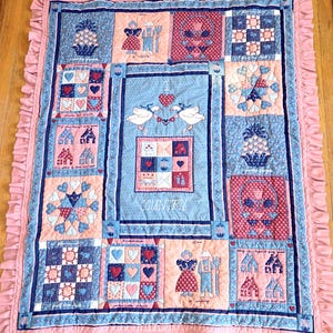 May include: A patchwork quilt with a light blue, pink, and navy color scheme. The quilt features various squares with heart, house, and floral patterns. The word "COUNTRY" is visible on the quilt. The quilt has a pink ruffled border.