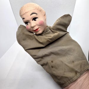 May include: A vintage hand puppet featuring a painted face with rosy cheeks, black eyes, and red lips. The puppet's head is attached to a tan fabric glove, showing signs of wear and age. The puppet's face has a neutral expression.
