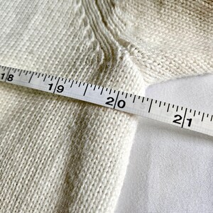 May include: Close-up of a cream-coloured knitted fabric with a measuring tape across it. The tape reads from 45.7 cm to 53.3 cm. The fabric has a textured appearance, and the background is a plain white surface.