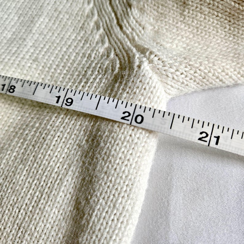 May include: Close-up of a cream-coloured knitted fabric with a measuring tape across it. The tape reads from 45.7 cm to 53.3 cm. The fabric has a textured appearance, and the background is a plain white surface.