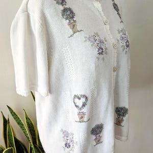 May include: A cream-coloured short-sleeved cardigan with embroidered floral and topiary designs in purple and green. The button-down sweater has a textured knit pattern and a scalloped neckline.