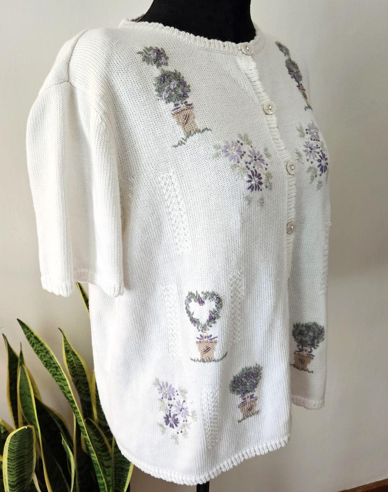 May include: A cream-coloured short-sleeved cardigan with embroidered floral and topiary designs in purple and green. The button-down sweater has a textured knit pattern and a scalloped neckline.