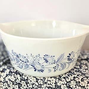 May include: A white mixing bowl with a blue floral design. The bowl has two handles and a textured exterior. The floral pattern includes stylized flowers and leaves. The bowl is likely made of ceramic or glass.