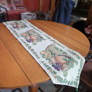 May include: Cream-colored table runner with a repeating pattern of a woven basket overflowing with fruit. The runner is bordered with green vines and floral accents, adding a touch of rustic charm.