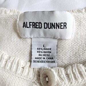 May include: Close-up of a cream-coloured knit sweater label with the text "ALFRED DUNNER". The label also includes size, fabric composition, RN number, and "MADE IN CHINA". A button is visible below the label.