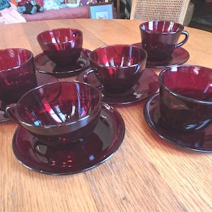 May include: A set of six ruby red glass teacups and saucers. The cups have a rounded shape with a handle, and the saucers are circular. The set is arranged on a wooden table, showcasing the rich color and design of the glassware.