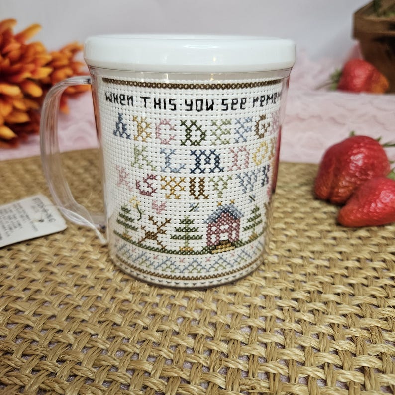 May include: A clear plastic mug with a white lid. The mug has a cross-stitch design on the side that says 'When this you see, remember me'. The design also includes a house, trees, and letters of the alphabet in various colors.