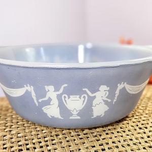 May include: A light blue ceramic bowl with a white design of two figures holding a banner and a vase. The bowl is on a woven surface.