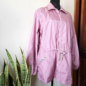 May include: A pink lightweight jacket with a zipper closure and a drawstring waist. The jacket has a floral embroidered design on the left pocket.