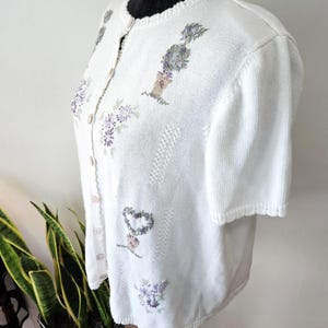 May include: White short-sleeved cardigan with embroidered floral and heart designs in shades of purple and green. The button-up sweater has a round neckline and a textured knit pattern. The cardigan is displayed on a black mannequin.
