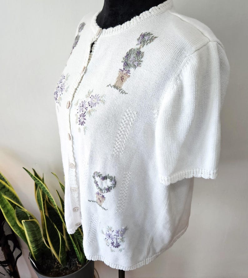 May include: White short-sleeved cardigan with embroidered floral and heart designs in shades of purple and green. The button-up sweater has a round neckline and a textured knit pattern. The cardigan is displayed on a black mannequin.