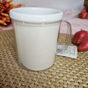 Pu&ograve; includere: Una tazza di plastica bianca con un manico in plastica trasparente e un inserto in rete bianca. La tazza &egrave; appoggiata su un tappetino intrecciato marrone.