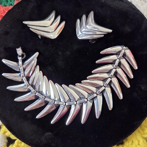 May include: A silver-toned jewelry set featuring a necklace and matching earrings. The necklace has a curved design with pointed, leaf-like elements. The earrings are clip-ons with a similar angular shape. The set is displayed on a black velvet surface.