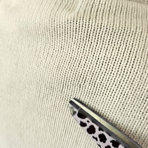 May include: Close-up of a cream-coloured knitted fabric with a textured surface. The fabric is in focus, with visible vertical and horizontal stitch patterns. A pair of scissors with a black and white spotted pattern on the handle is in the lower right corner.