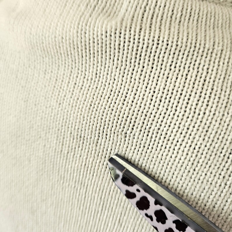 May include: Close-up of a cream-coloured knitted fabric with a textured surface. The fabric is in focus, with visible vertical and horizontal stitch patterns. A pair of scissors with a black and white spotted pattern on the handle is in the lower right corner.