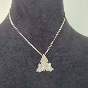 May include: Silver-toned pendant necklace featuring a detailed, geometric design. The pendant is suspended from a delicate chain. The necklace is displayed against a dark background, highlighting the metallic sheen of the jewelry.