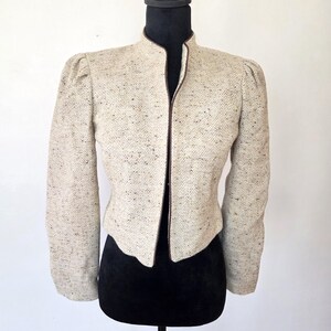 May include: A beige tweed jacket with a stand-up collar and long sleeves. The jacket has a brown trim along the collar and front edges. The jacket is displayed on a black mannequin. The fabric has a subtle herringbone pattern.