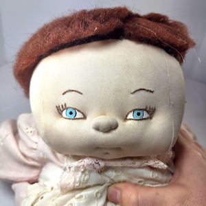 May include: A soft fabric doll with a round, cream-colored face, short brown yarn hair, and bright blue embroidered eyes. The doll features a small, rounded nose and stitched eyebrows, wearing a pale pink dress with white eyelet lace trim.