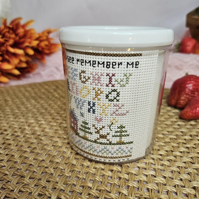 Pu&ograve; includere: Un contenitore di plastica bianco con un inserto di vetro trasparente. L'inserto di vetro ha uno sfondo bianco con un motivo a punto croce colorato che dice "See Remember Me".
