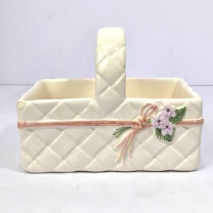 May include: Cream-colored ceramic basket with a woven pattern, a handle, and a pink ribbon accent. The basket is decorated with a small bouquet of purple flowers and green leaves. A decorative piece for the home.